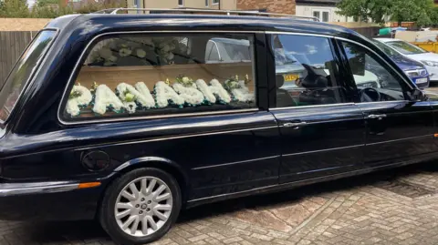 A black hearse with a pale wood coffin inside, with a floral display saying "Panashe" in front of it.