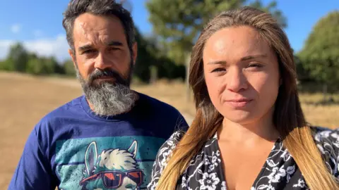 A man and woman sitting one slightly behind the other. The man has a greying beard and is wearing a blue T shirt. The woman has long brown hair wearing a black and white patterned dress 