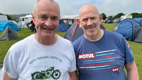 Paddy Corey and John Little stand in field of tents. Paddy wears a grey t-shirt with a green motorcycle on it, while John wears a blue T-shirt with white, blue and red stripes.