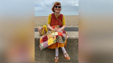 Contributed Marcia Riddington with red hair sits on a sea wall smiling at the camera. She wearing a yellow top with a red vest over the top and a skirt made up of a former quilt with different patches of fabric.