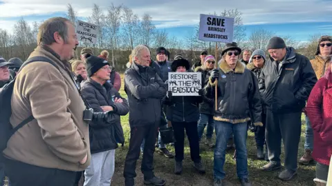 A group of about 20 people, some with placards about saving the headstocks, listen to a speaker