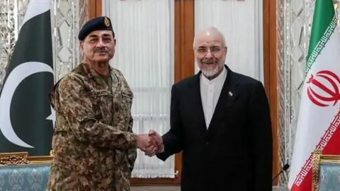 Reuters Iran's parliament speaker wearing a dark jacket over a white shirt shakes hands with Pakistan's army chief who's wearing military fatigues and a green beret. Both men are smiling, and the flags of both countries can be seen alongside each on the edges of the image.