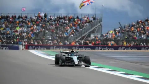 EPA A Formula 1 car on the Silverstone circuit. The car is green, black and silver. In the background is a grandstand full of fans looking on.