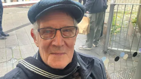  Alan Benison smiles wearing a flat cap outside while waiting for the new bus. 