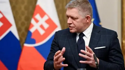 Reuters Robert Fico talks to reporters, wearing a dark suit and tie with a backdrop of two Slovak flags