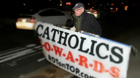 BBC John holding a sign saying "Catholics Cowards" on the road outside the Vatican Embassy