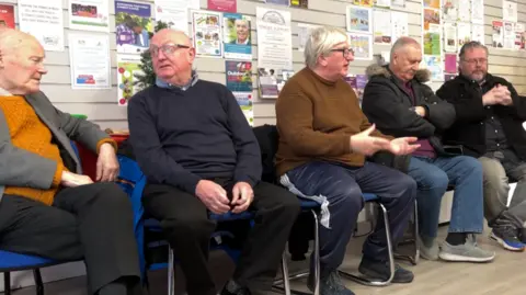 A group of men are pictured seated in a row in a community centre in Blyth in Northumberland. They are all in conversation and are all over the age of 40. 