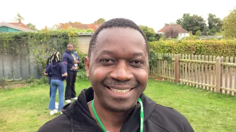 Luke Deal/BBC Herbert Mashami is standing outside on some grass and is smiling into the camera. He has short black hair and is wearing a green lanyard. He is wearing a black jacket and behind him are a group of children singing while standing in a circle.