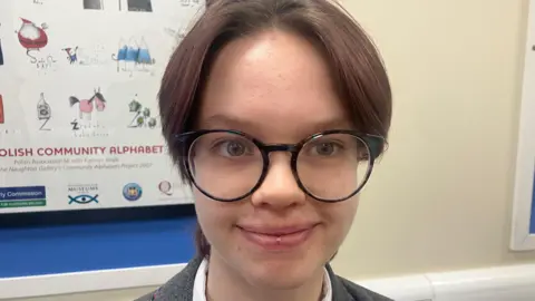 Olivia is smiling at the camera. She has short brown hair and black framed glasses. She is wearing a white shirt and grey blazer. 