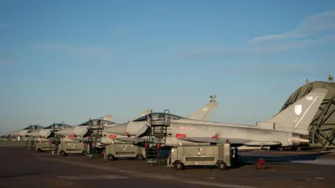 Grey typhoon jets with are mounted onto small four-wheeled units with steps leading up to each one in a row. They are located on a base and there are four of them.
