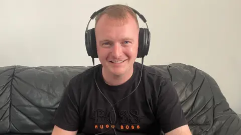A man wearing large black headphones and a black t-shirt, while sitting on a black leather sofa.
