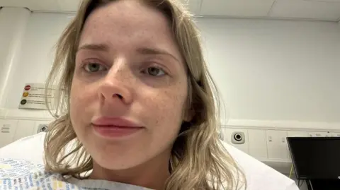 Close-up of Ellie lying in a hospital bed, wearing a patterned hospital gown, with medical wall equipment and a monitor visible in the background.