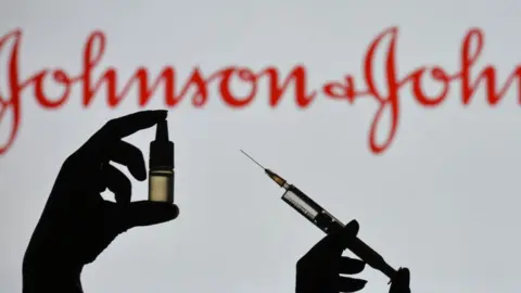 Getty Images Silhouette of a man holding a medical syringe and a vial seen displayed in front of the Johnson and Johnson logo on a screen