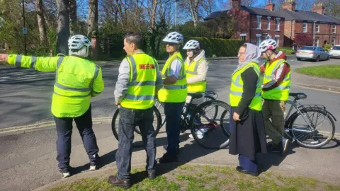 A group of people wearing hi-vis jackets over their usual clothes, some holding up bikes, taking place in the scheme on a quiet road with few cars. 
