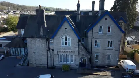 Aerial view of the building that used to be St Monicas. A large stone building with steep pitched roofs and blue-trimmed gables, featuring multiple chimneys and adjoining extensions, set within a residential area with houses in the background.