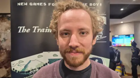 A young man with curly hair and a beard wearing a purple jumper infront of a banner that says "new games for Game Boy!"