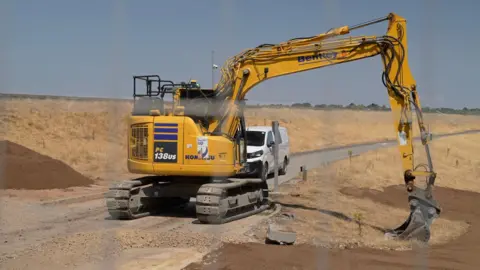 A yellow digger parked beside a road on a dry embankment, with a white van and piles of earth nearby.