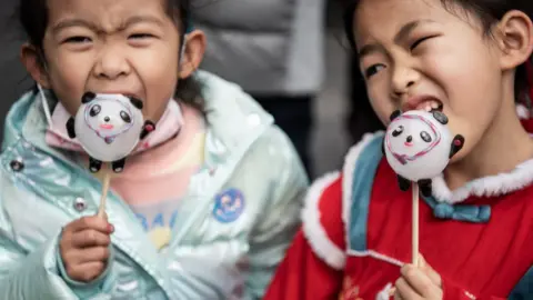 Getty Images Two girls lick sugar figurine in the likes of Beijing 2022 Winter Olympic mascot Bing Dwen Dwen during the celebration of the Lantern Festival which marks the end of Lunar New Year celebrations.