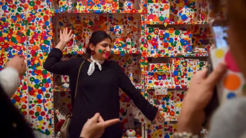 Getty Images A gallery visitor poses in front of a wall covered in multi-coloured spots on the final day of the Yayoi Kusama: Infinity Mirrors exhibit at the Smithsonian Institute's Hirshhorn Museum on Sunday, May 14, 2017