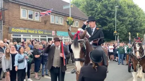 Man in top hat on horseback reads a proclamation near shops