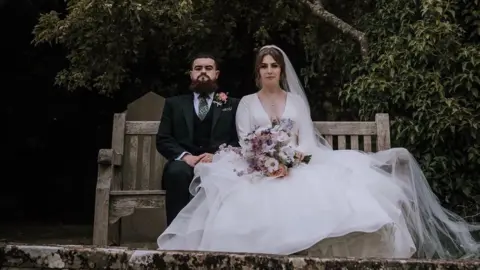 A bride and groom sit side-by-side on a wooden bench looking at the camera, with expressionless faces. The groom is wearing a dark suit with a pink flower attached to his jacket. The bride is wearing a long white dress draped over the side of the bench, and is holding a bouquet of flowers. 