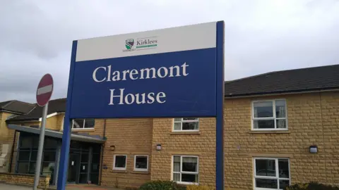 Image shows a two storey sandstone building with white framed windows. 
In the foreground is a blue and white sign with 'Claremont House' on which is the name of the home. 