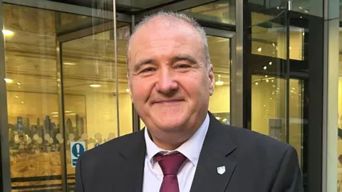 Lee Castleton has receding grey hair and is wearing a black blazer over a white shirt and burgundy tie. He is smiling while standing in front of a glass building.