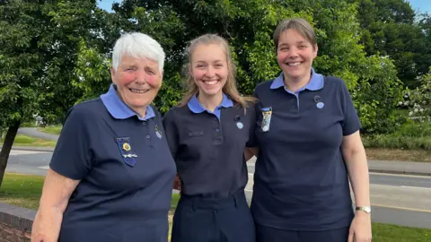 (L-R) Gillian Watson, Megan Megahey and Hazel Megahey. They are standing outside, wearing Girlguiding uniforms. They have their arms around each other and they are all smiling. There is grass and trees in the background and it is sunny.