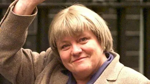 Getty Image Northern Ireland Secretary, Mo Mowlam, arrives at No 10 Downing Street. She has short blond hair. She's waving and wearing a tweed jacket or coat.