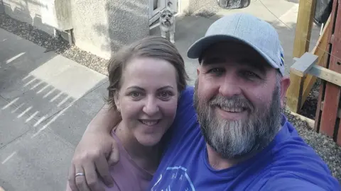 A man with a longer beard is standing in front of a house with his arm around a woman with short blond hair. She is wearing a pink top, while he is dressed in a dark-blue t-shirt and a grey cap. There is a schnauzer dog visible behind the couple.