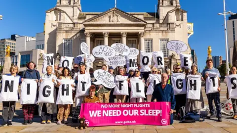 GALBA Protestors holding white pillows with the sentence 'No More Night Flights' spelled out in black letters on them. They are stood in front of Leeds Civic Hall 