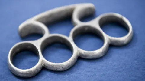 A silver metal knuckleduster. It is a fitted weapon designed to be worn around the knuckles of the hand.