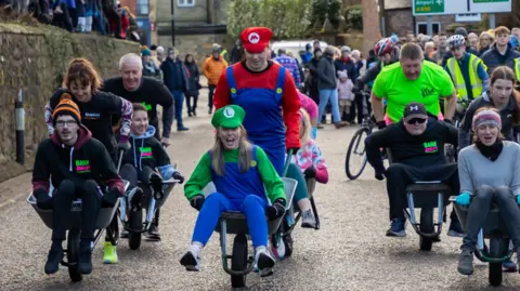 People, some of them in fancy dress, are being pushed in wheelbarrows down a road. Crowds are lining the road.
