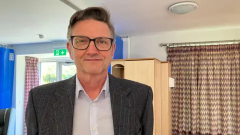 Jeremy Lune is wearing a suit and stands in the foreground of a room with cupboards, curtains and a glass door with an emergency exit sign above.