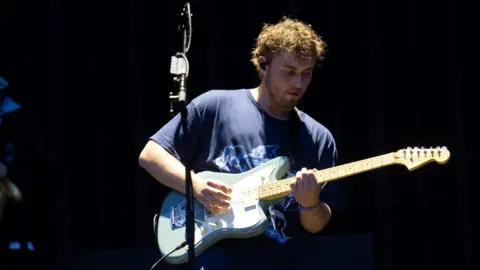 Getty Images Sam Fender on stage, playing a blue and white electric guitar. He wears a blue T-shirt and the staging behind him is dark.