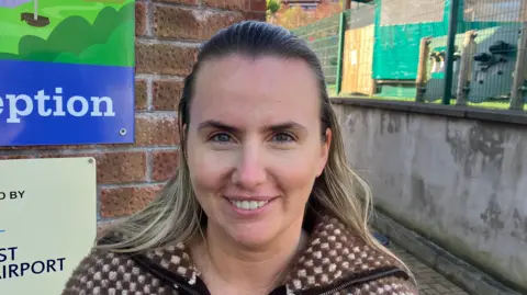 Rodgers has long brown hair which is highlighted blonde. She is wearing a brown checked fleece.