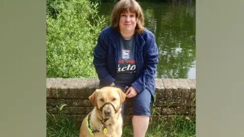 Zoe Newson Lisa Newson, dressed in navy blue cropped trousers, an Ipswich Town t-shirt and navy blue jacket sits on a small brick wall. In front of her is her yellow coloured guide dog Holly.