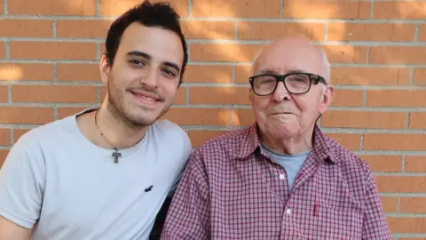 Grandson and grandfather sit on a bench in a sun-dappled view against a brick wall