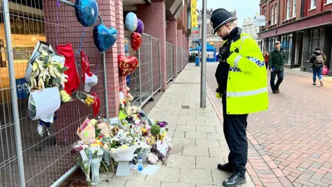 John Fairhall/BBC A policeman looking at the flowers left in tribute to James Quigley
