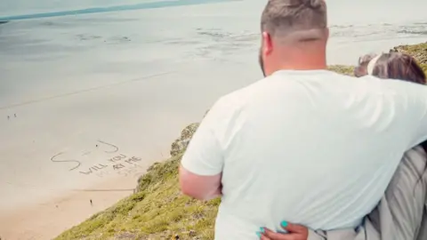 @d0oner The couple look down at the proposal message written in the sand below