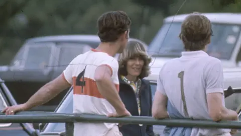 Shutterstock Charles stands next to Camilla, near parked cars on the edge of a polo match