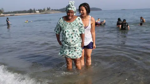 Getty Images A Congolese woman stands in the ocean for the first time guided by an American woman during during a programme to help integrate new refugees in Maine, the US - Tuesday 28 August 2018