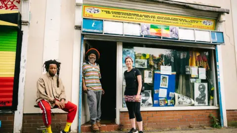 Khali Ackford Two people standing and one sitting in front of the Rastafarian Centre, which has a yellow sign