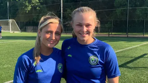 Zoe (left), and Kelly (right) in Sion Swift's navy blue training t-shirts, which bears the club's badge - a white circle with a football in the middle. Over the top of the football is the lime green outline of a swift - a swallow-like bired. Zoe and Kelly are on the team's pitch, which looks well-kept and is a healthy green colour. Both are smiling, and Kelly's arm is around Zoe's shoulder.