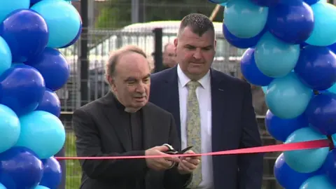 Francis Lagan looks on as a priest cuts a ribbon to open a new sports pitch at St Mary's Primary School