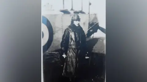 Hertfordshire Archives and Local Studies A black and white image of the female aviator wearing a long leather coat and flight head gear, standing in front of a propeller plane
