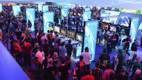 Getty Images The showfloor of a gaming expo viewed from an elevated angle. It's very crowded, and there are rows of TV screens hooked up to consoles. People can be seen playing a range of different games. The scene is dotted with tall, brightly light boards displaying the PS3 logo