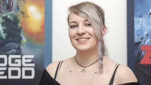 A smiling young woman with dyed silver hair, in an asymmetric style with a long, ponytail-style fringe to one side. She's wearing a black, off-the shoulder top and a necklace on a short black string with a silver pendant hanging from it. The pendant design is a gaming joypad mixed with the seated stick figure more commonly seen with a wheelchair on disabled signage. Behind her two pictures hang on the wall - one shows comic book character Judge Dredd and the other a zombie soldier with yellow, glowing eyes wearing a tattered military helmet.