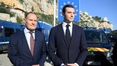 NICOLAS TUCAT/AFP French far-right Rassemblement National (RN) party President Jordan Bardella (R) and RN party member and former head of the EU's border agency Frontex Fabrice Leggeri (L) look on, with a sign reading "Italy" and French gendarmerie vehicles in the background, during a visit at a border crossing between France and Italy in Menton, southeastern France, on February 19, 2024