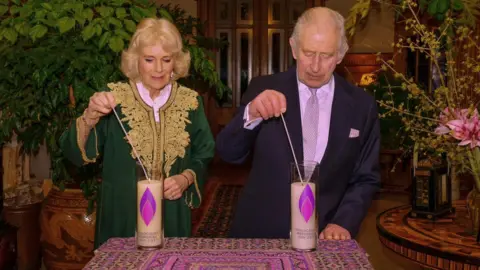 Royal Family Camilla and Charles stand behind a table on which two large candles decorated with a purple flame. They each hold a long match to the wick.